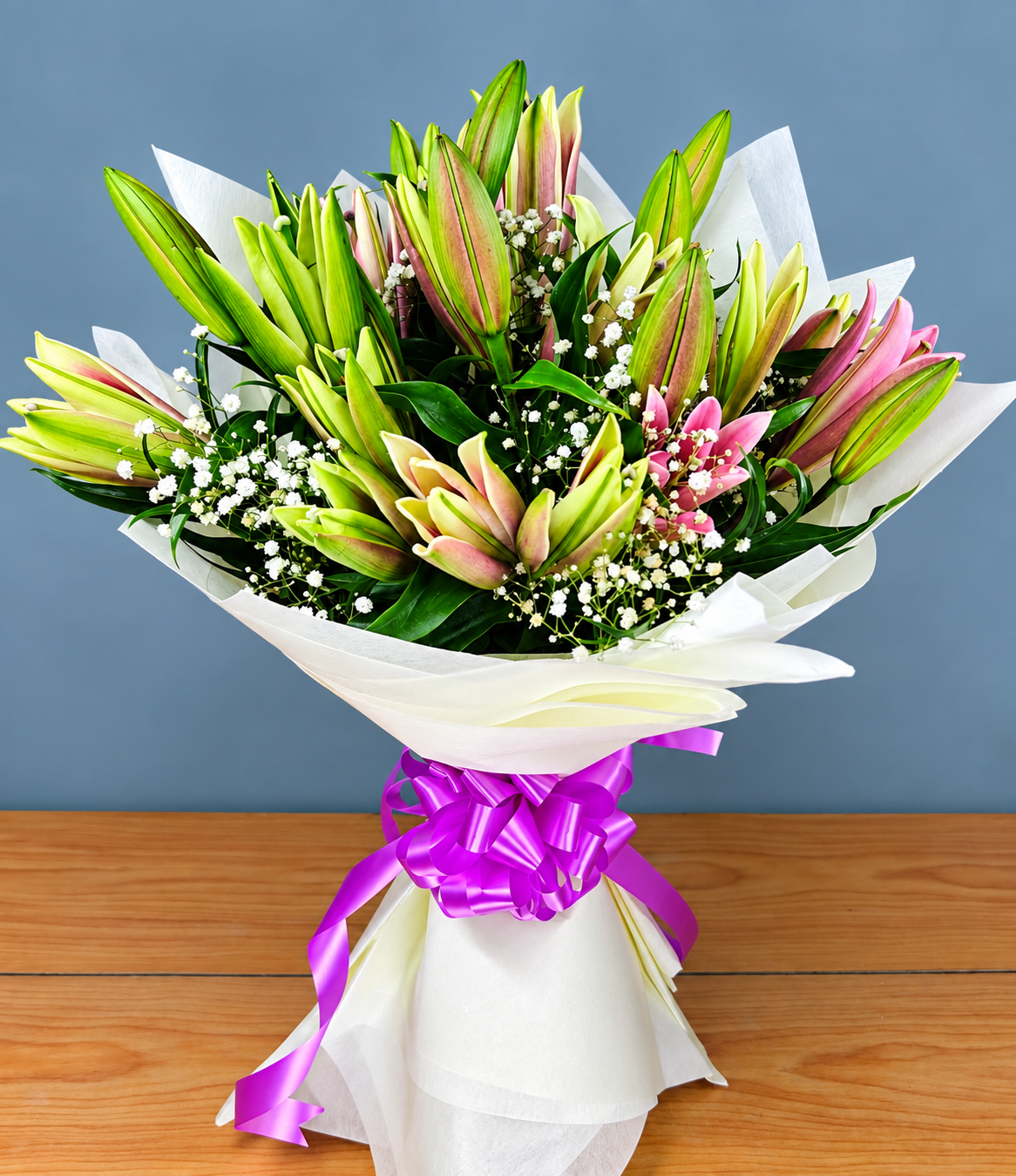 Lily Bloom Wrap bouquet with pink-tinted lily buds, white baby’s breath, fresh greenery and purple ribbon by Whitefields
